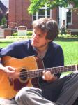 photo of Jon Watts sitting on the grass on a sunny day, playing guitar and singing