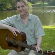 photo of Ben Tousley sitting by a lake and holding his guitar on his lap.