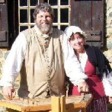 photo of John and Jan Haigis standing outside by a split-rail fence on a sunny day, wearing pioneer costumes